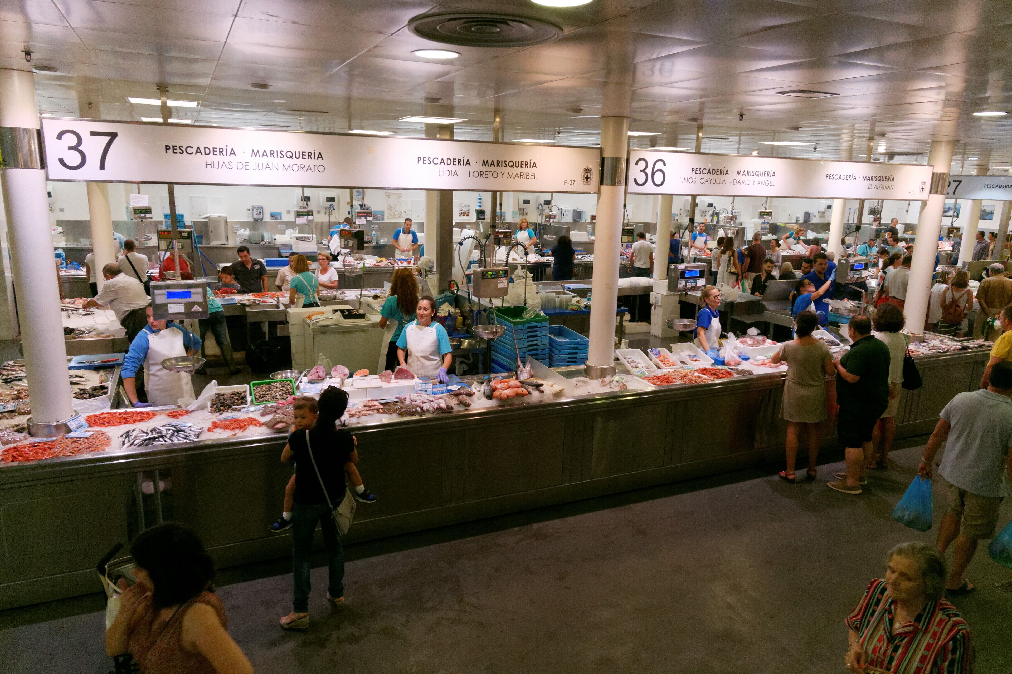 Een levendige vismarkt met meerdere kraampjes, elk aangeduid met een nummer, waar mensen zeevruchten uitkiezen.