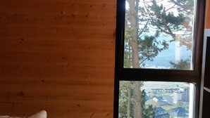 A wooden cabin room with a bed, a large window, and a view of trees and buildings outside.
