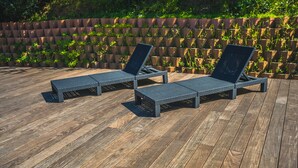 Dos sillones reclinables negros sobre una terraza de madera con un muro de ladrillos como fondo.
