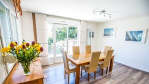 Un comedor con una mesa de vidrio y sillas de mimbre, un jarrón de flores sobre una mesita de madera, y vistas al jardín a través de puertas de cristal.