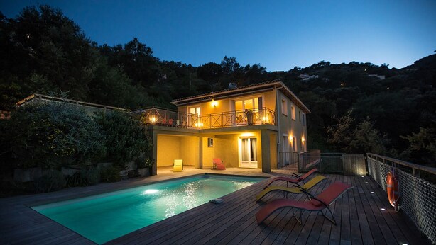 Una casa de dos pisos con piscina, terraza de madera y mobiliario exterior.
