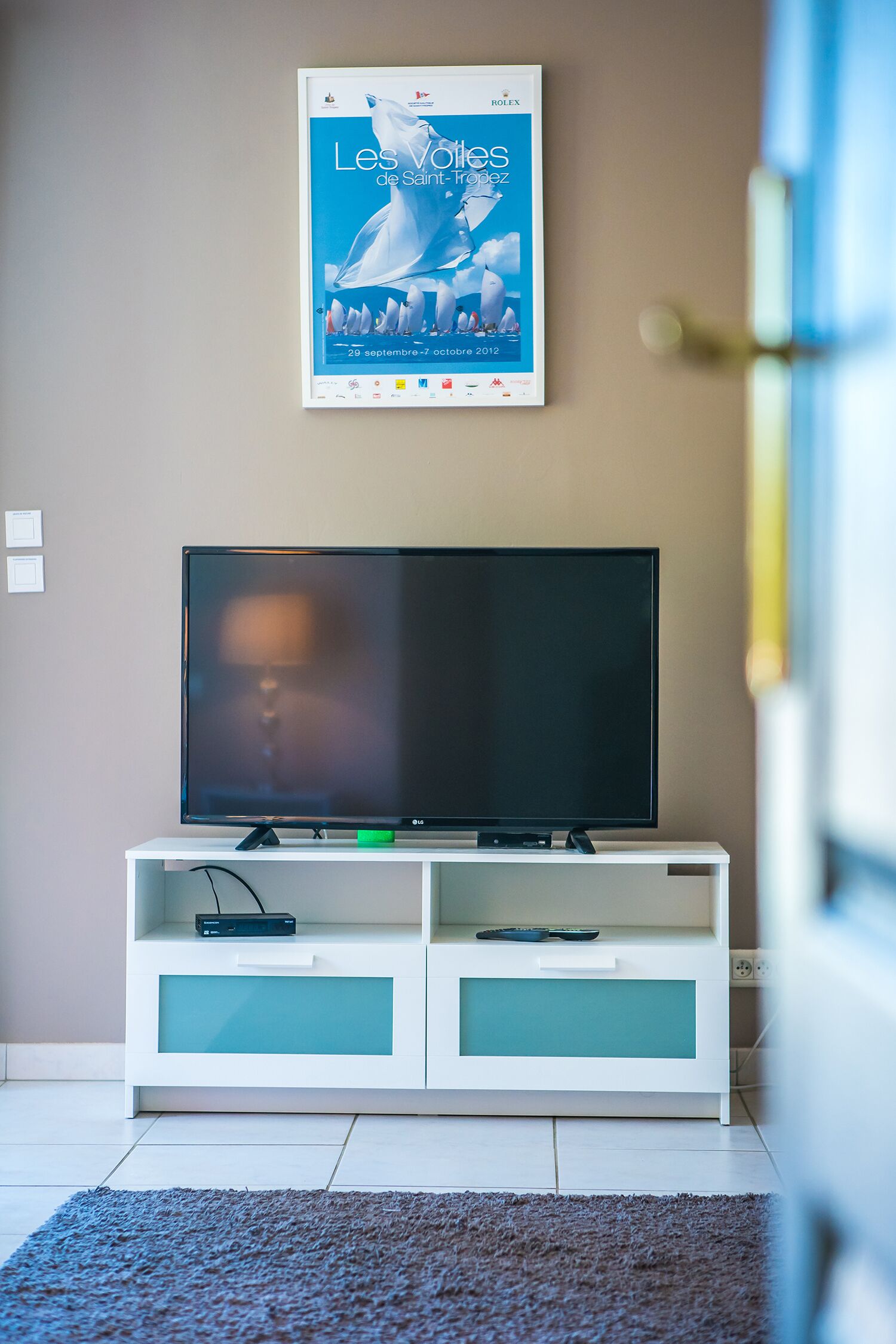 A living room with a flat-screen TV on a white entertainment unit, a poster on the wall, and a door to the right.