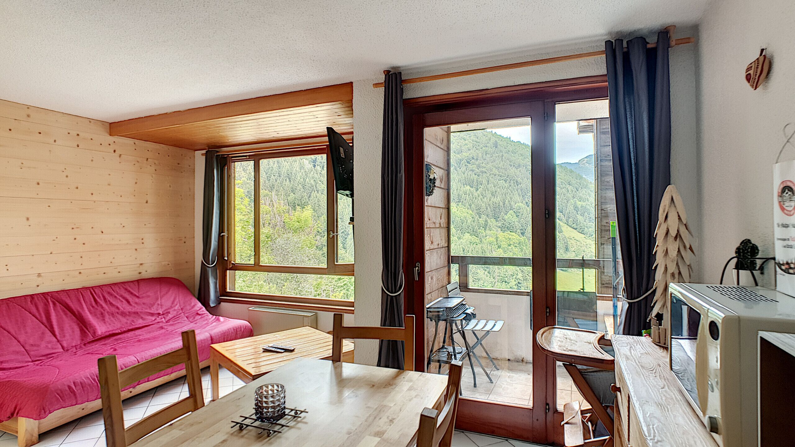 Una habitación acogedora con suelo de madera, un sofá rojo, una mesa de madera y un escritorio con una computadora. Amplias ventanas ofrecen vistas a un área boscosa.