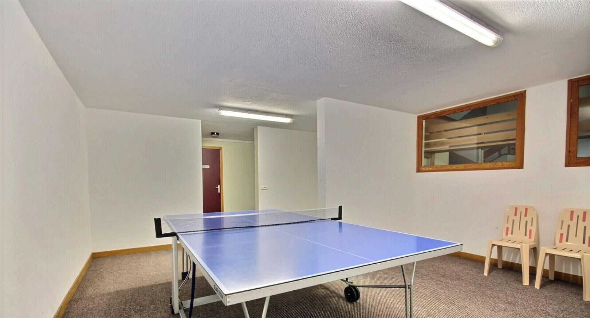 A room with a ping pong table, carpeted floor, two wooden chairs, and a door in the background.