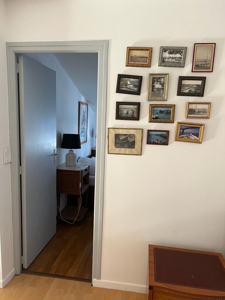 Ein Zimmer mit einer Tür zu einem Flur, einem Nachttisch mit Lampe und einer Bildergalerie an der Wand.