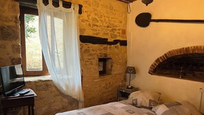 A rustic bedroom with stone walls, a bed with patterned bedding, a television on a wooden table, and a fireplace. - Gîte 2p Lascaux, 2*, wifi, park, games, heated swimming pool, Spa, ok dogs & cats (MONTIGNAC-LASCAUX)