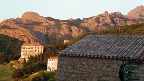 Un bâtiment en pierre avec un toit recouvert de tuiles et une enseigne indiquant un restaurant. En arrière-plan, on aperçoit deux immeubles à plusieurs étages, coiffés de toits rouges, qui se dressent sur un paysage montagneux.
