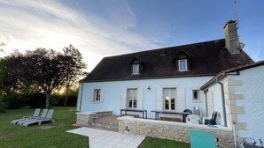 A two-story house with a white exterior, a dark roof, and a chimney. There is a patio with outdoor furniture and a small garden area.