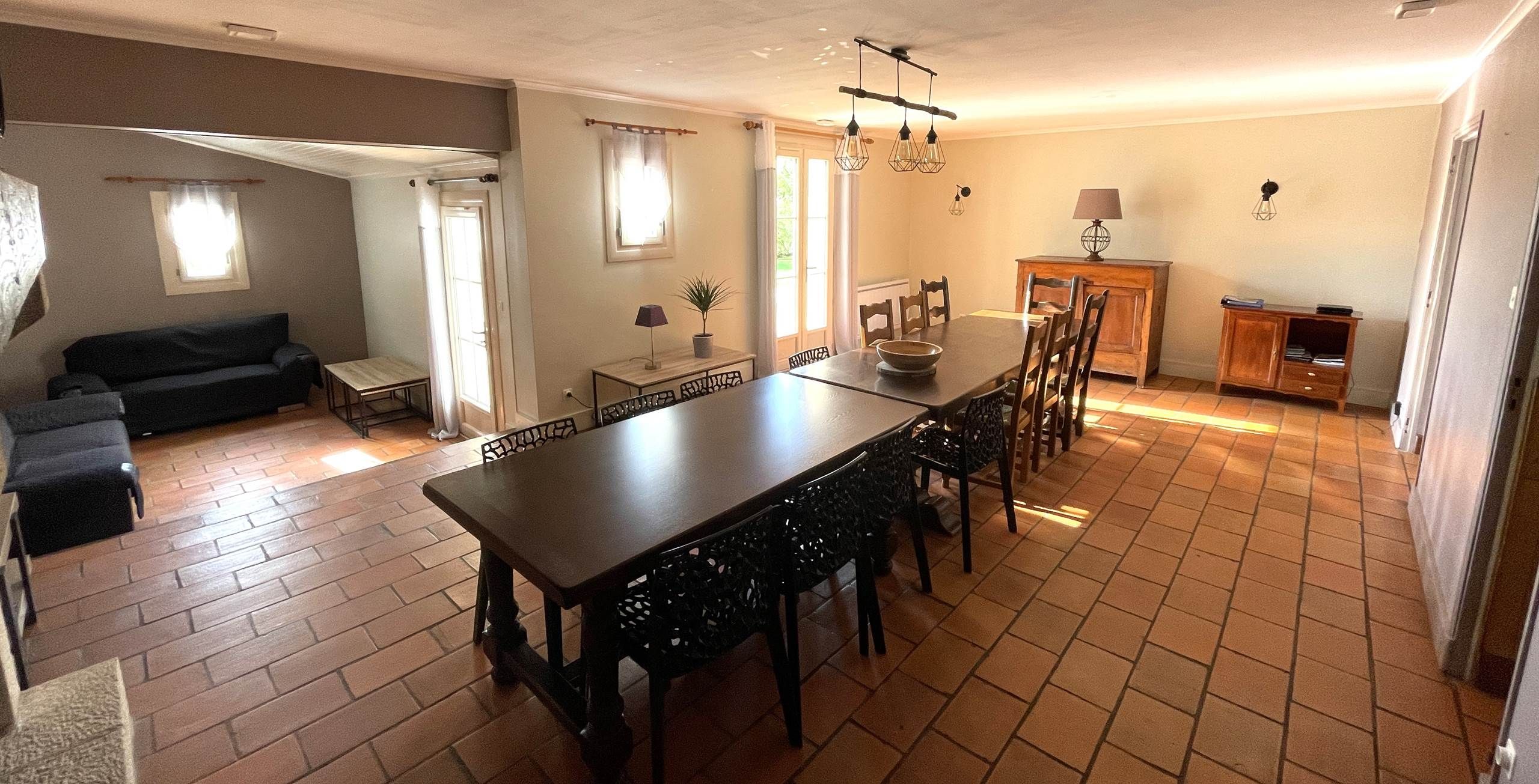 Un espace repas comprenant une longue table et des chaises, un canapé, une petite table avec une lampe et un buffet.
