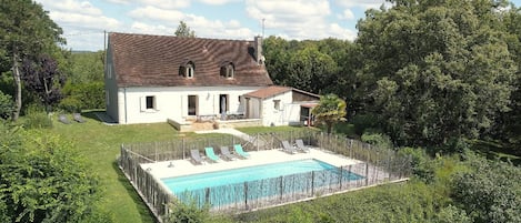 A house with a swimming pool surrounded by a wooden fence, a garden, and a large tree.
