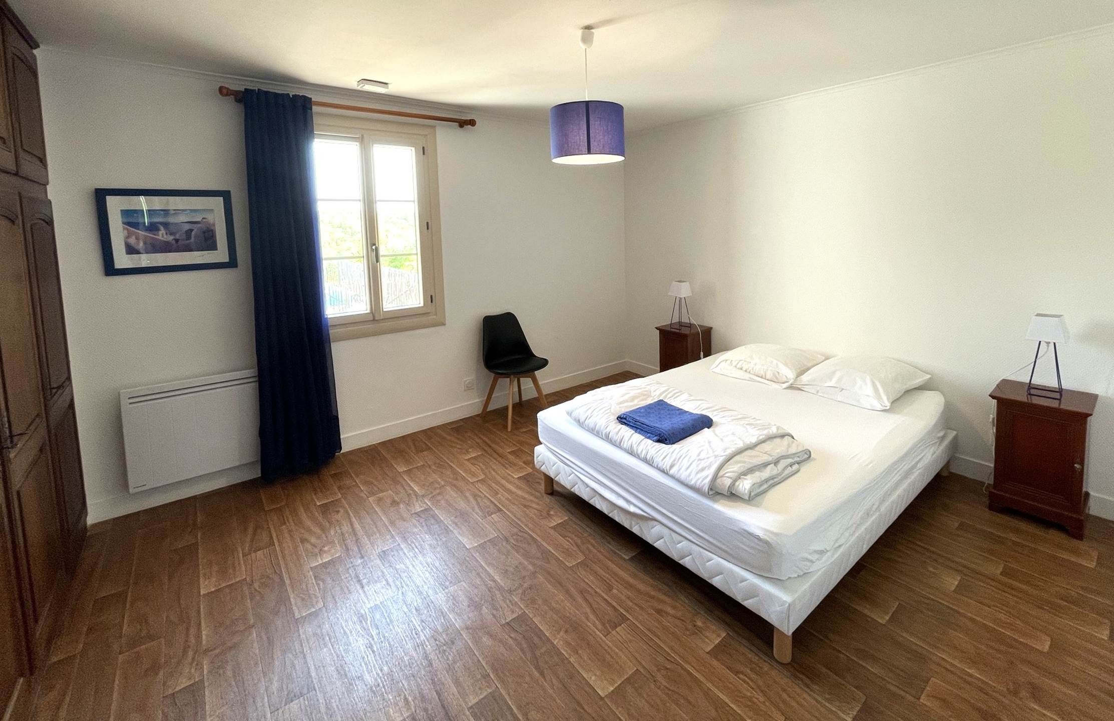 A bedroom with a bed, a chair, a bedside table, a lamp, a window with curtains, and a picture on the wall.