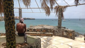 Un hombre sentado en un banco de piedra contemplando una playa con barcos a lo lejos.