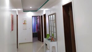 Un pasillo con paredes blancas, techo blanco con detalles verdes y luces empotradas. A la derecha, hay puertas, una consola blanca con un espejo y objetos decorativos, y un cartel en la pared izquierda.