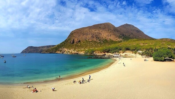 Una playa con agua turquesa cristalina, orilla de arena y una montaña al fondo.