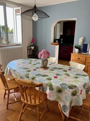 A dining area with a round table, wooden chairs, a floral tablecloth, a vase with flowers, and a kitchen in the background.