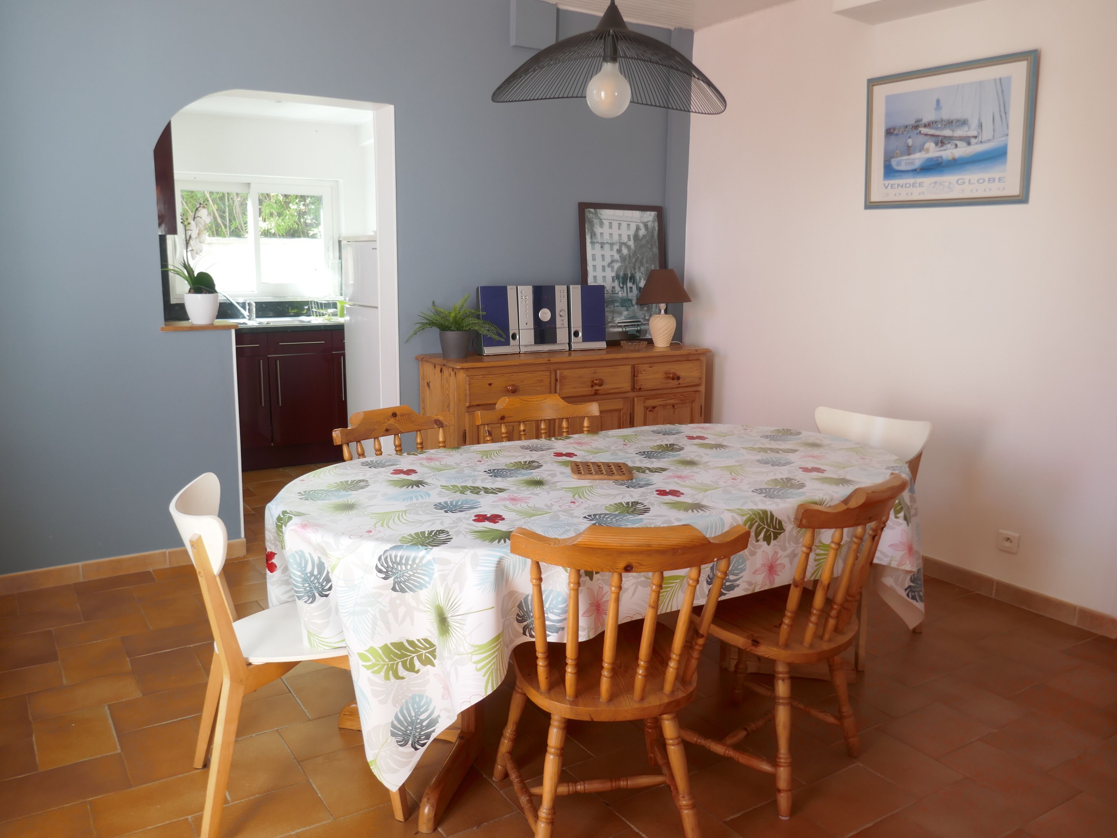 A dining area with a table set for four, surrounded by wooden chairs. A cabinet with drawers and a television is against the wall.
