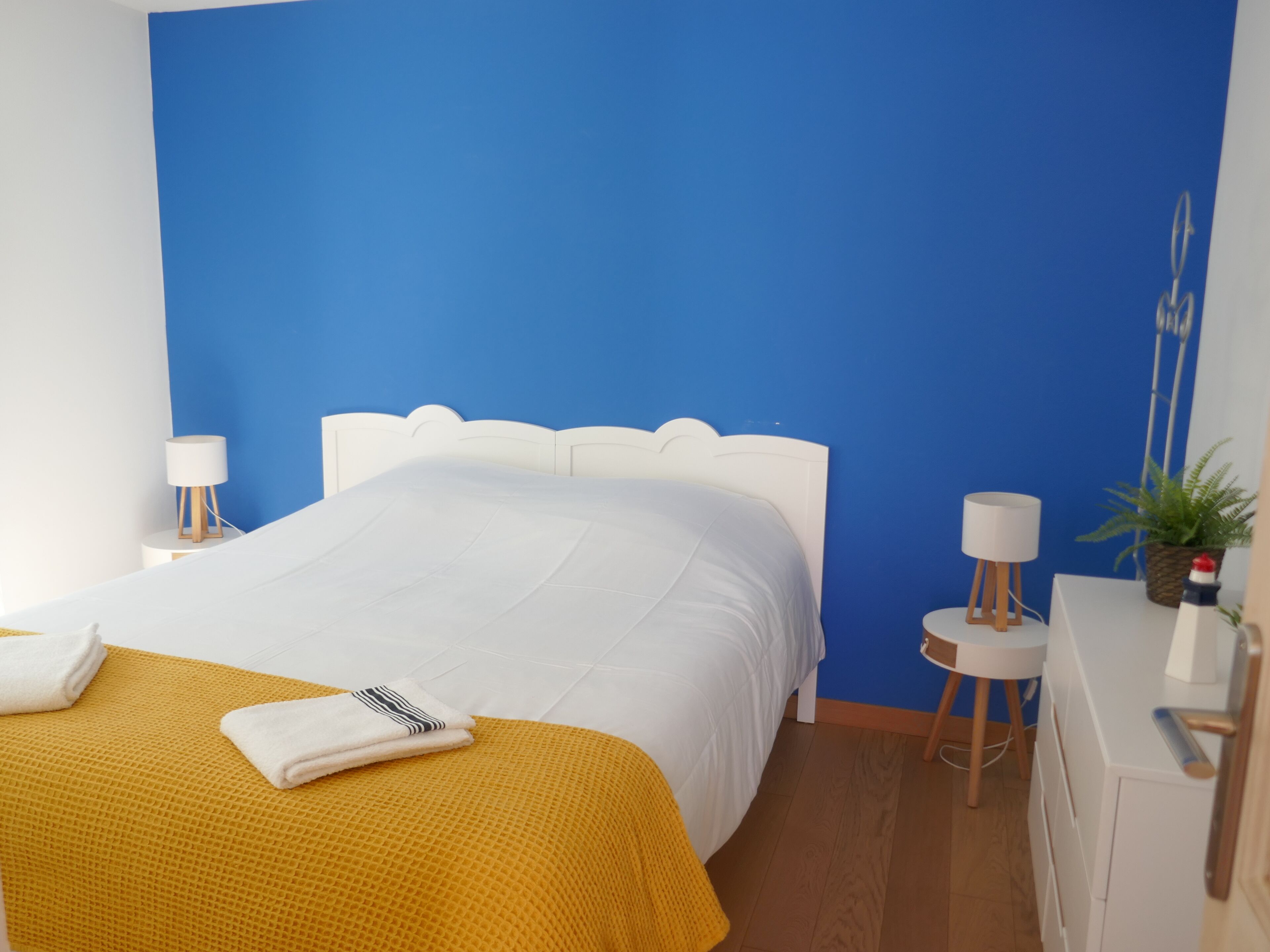 Een net opgemaakt bed met een witte deken en een mosterdgele plaid, een wit hoofdbord, twee nachtkastjes met lampen, een kleine plant en een houten dressoir.