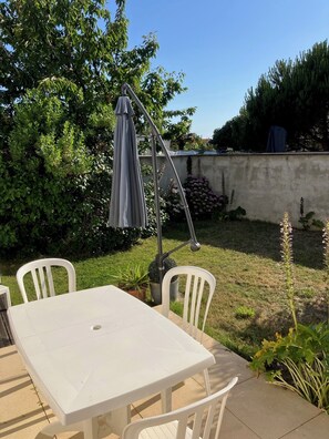 A patio with a white table and chairs, a gray umbrella, and a garden with various plants and a wall in the background.