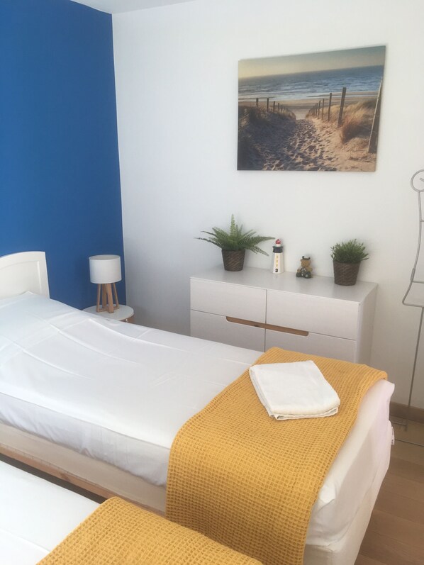 A bedroom with a bed, a bedside table, a lamp, a dresser, and a painting of a beach scene.