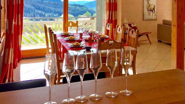 Un coin repas avec une table dressée pour six personnes, de grandes fenêtres offrant une vue sur les montagnes et un tableau représentant un cerf accroché au mur.