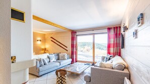 Un salon chaleureux avec un canapé, une table basse et une cheminée. Des paires de skis sont appuyées contre le mur, et il y a une fenêtre avec des rideaux.