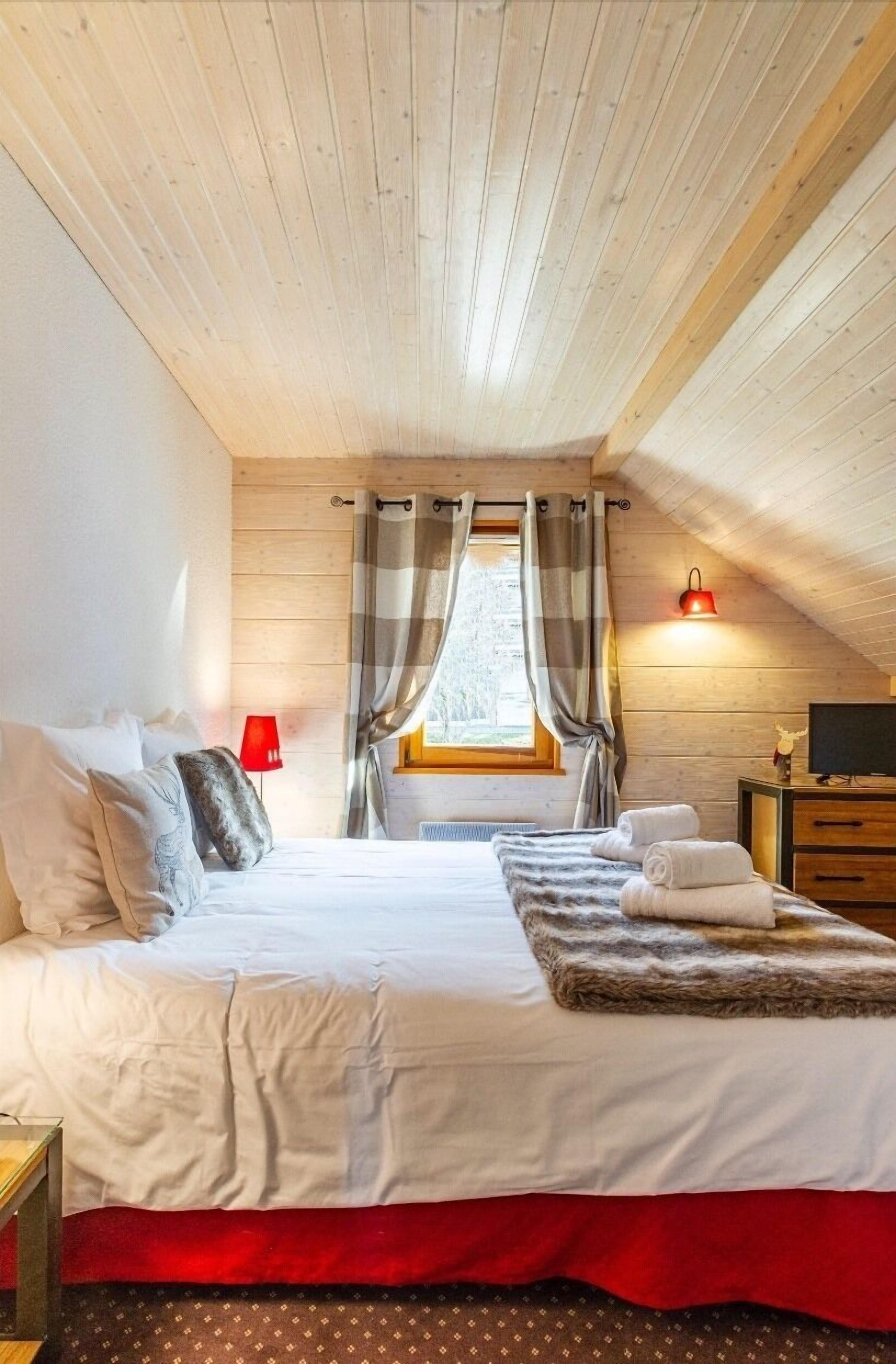 Une chambre confortable avec un plafond en bois, un grand lit recouvert de draps blancs, un plaid rouge et une fenêtre avec des rideaux.