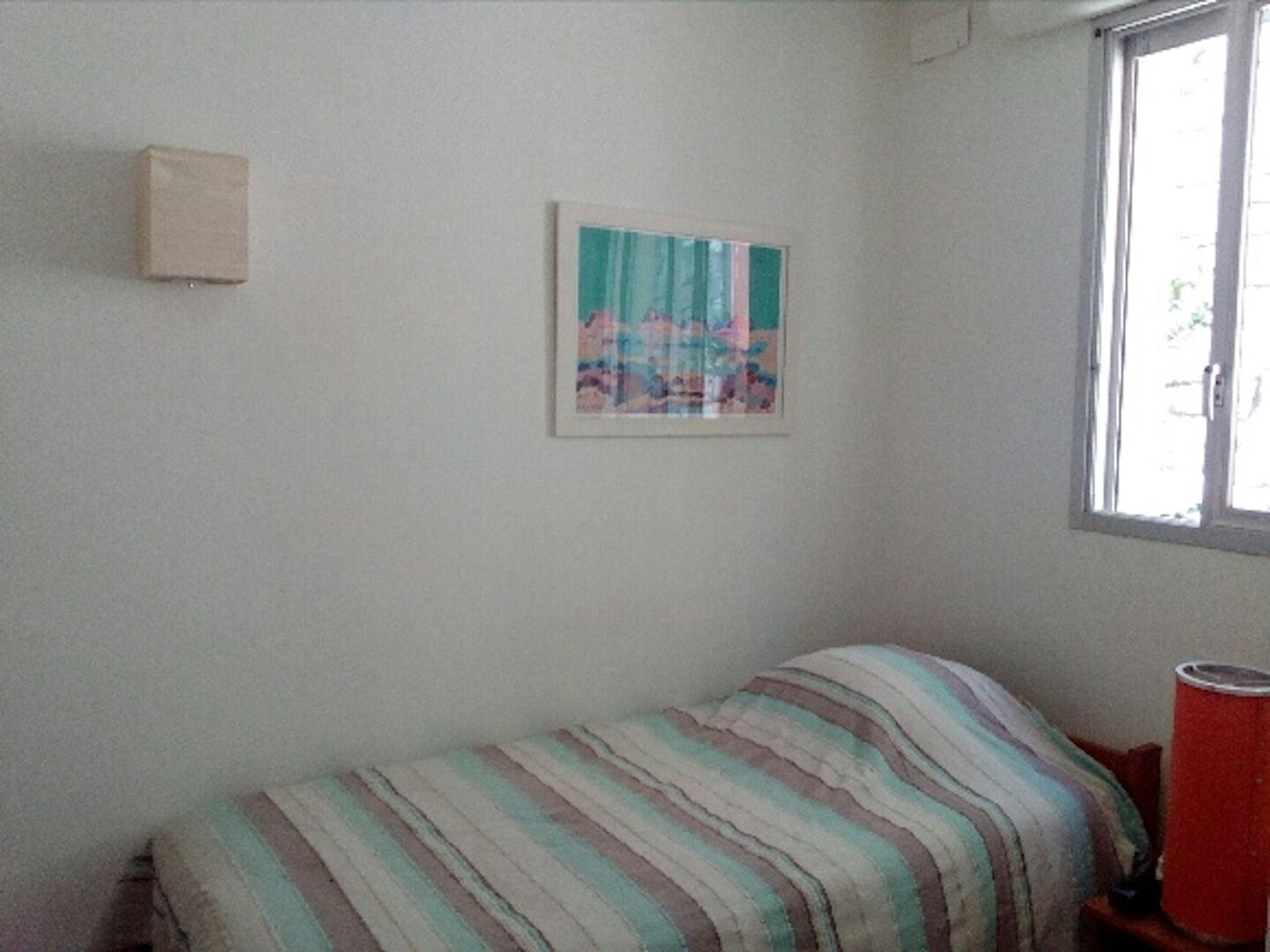 Una habitación pequeña con cama individual, una colcha a rayas, un cuadro en la pared y una ventana con un objeto rojo en el alféizar.