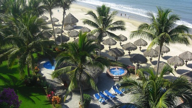 A beach resort with palm trees, thatched umbrellas, a pool, and a view of the ocean.