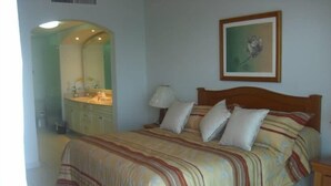 A bedroom with a bed, a nightstand, a lamp, a framed picture on the wall, and a bathroom visible through an archway.