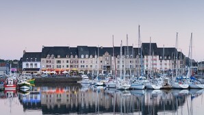 Una marina con varios barcos amarrados, edificios al fondo y una superficie acuática tranquila que refleja los barcos y edificios.