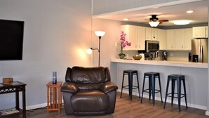 Un salon moderne avec un fauteuil en cuir, une petite table d’appoint en bois et un lampadaire. On aperçoit à l’arrière-plan un coin cuisine avec des meubles blancs et un tabouret de bar.