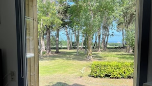 Ein Zimmer mit großem Fenster, das auf eine grüne Fläche mit Bäumen und Meerblick blickt.