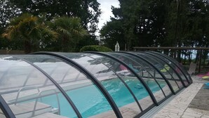 Ein überdachter Swimmingpool mit transparenter Dachkonstruktion, umgeben von Steinfliesen und Holzterrasse.