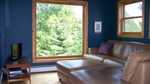 Ein Zimmer mit großem Fenster, einem braunen Ledersofa, einem Holztisch und einem Fernseher auf einem Schrank.