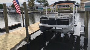 A pontoon boat docked at a wooden pier with an American flag, a water taxi, and a house in the background. - Waterfront Home on Canal - Heated Pool, Free use of Pontoon Boat! Discount's (New Port Richey)