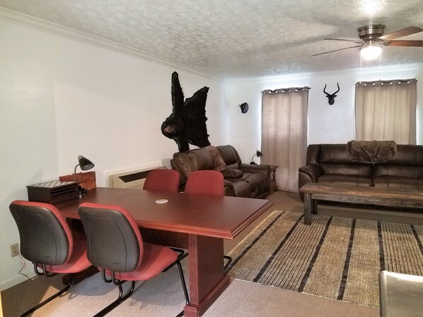 A living room with a dining table, red and black chairs, a brown leather sofa, a ceiling fan, and decorative wall mounts.