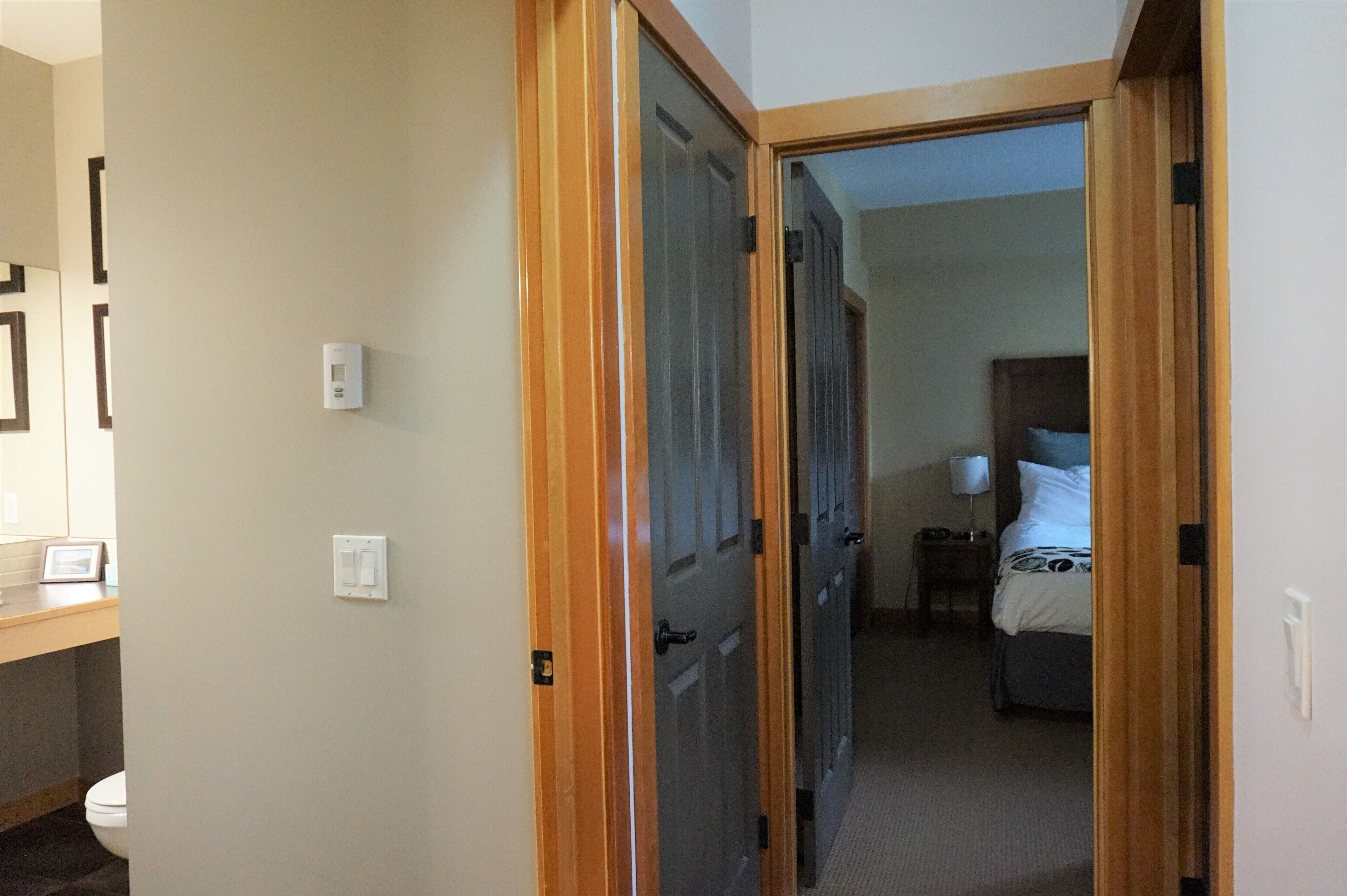 Habitación de hotel con puerta de madera, cama con edredón blanco, mesita de noche con lámpara y pequeño tocador.