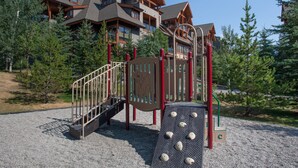 A playground with a climbing structure and a slide, surrounded by trees and a lodge in the background. - Premium 2 Bedroom, 2 Bath Condo w/central Air Conditioning- Lodges at Canmore (Canmore)