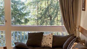 A cozy room with a wooden dining table, a cushioned sofa with patterned pillows, and a view of a forest through large windows.