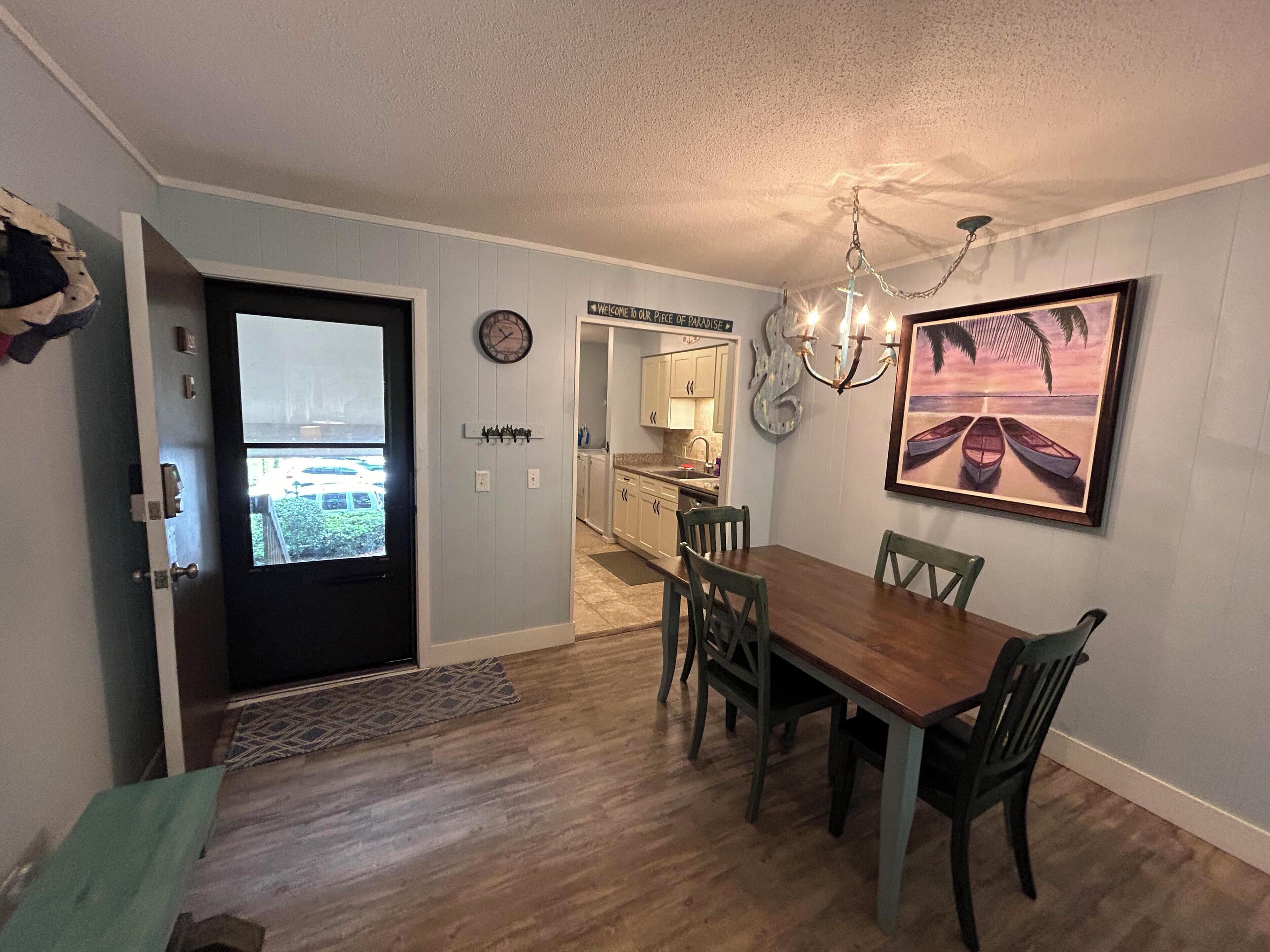 A dining area with a wooden table and chairs, a chandelier, a clock, and a framed picture of boats on a beach.