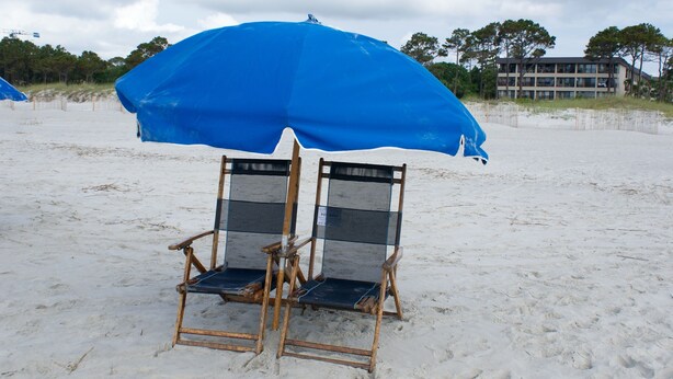 Zwei Strandstühle unter einem blauen Sonnenschirm an einem Sandstrand, im Hintergrund ein Gebäude.