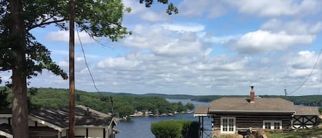 A rustic cabin with a dirt driveway, a wooden dock extending into a lake, and a backdrop of rolling hills and a partly cloudy sky.