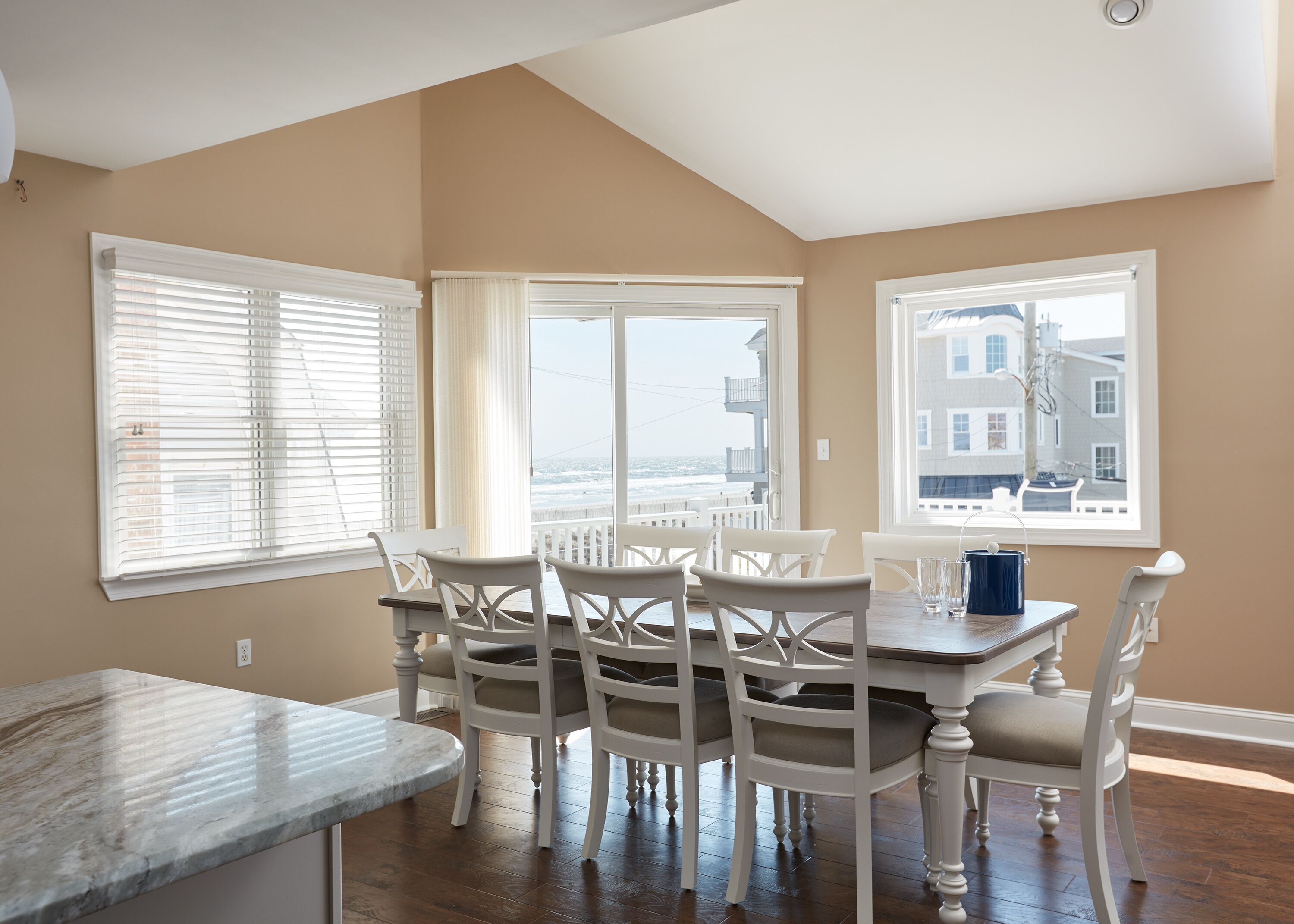 A dining area with a marble countertop, white chairs, and large windows offering a view of the ocean and buildings.