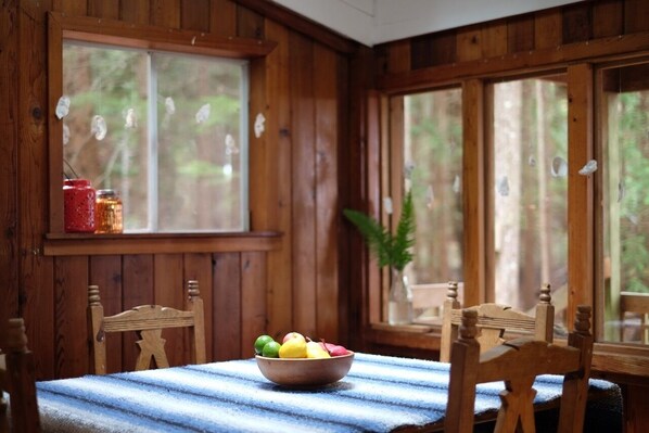A wooden cabin interior with a dining table, chairs, a bowl of fruit, and a window with a view of trees. - Off-Grid Island Hideaway | Walk to Stunning White Sand Beach (Savary Island)