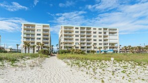 Ein Hotel direkt am Strand mit mehreren Etagen und Balkonen, ein sandiger Weg führt zum Eingang, und ein klarer, blauer Himmel.