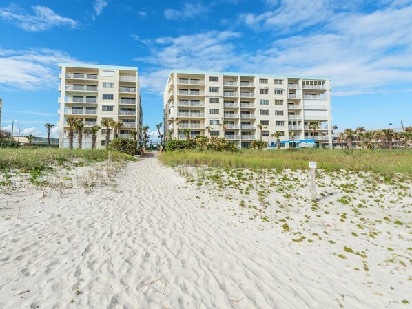 A beachfront hotel with multiple floors and balconies, a sandy path leading to the entrance, and a clear blue sky.