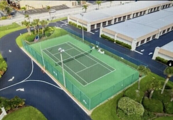 An aerial view of a tennis court surrounded by a green fence, adjacent to a building with a flat roof and a parking lot.