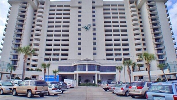 A large, multi-story hotel with a central entrance, numerous balconies, and a parking lot with several cars.