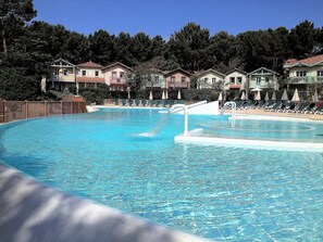 Una amplia piscina con múltiples carriles, rodeada de sillones de descanso y sombrillas, con villas al fondo.
