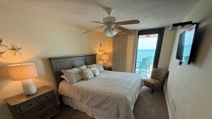 Quarto de hotel com cama, mesa de cabeceira com abajur, ventoinha no teto, cadeira e televisão fixada na parede.